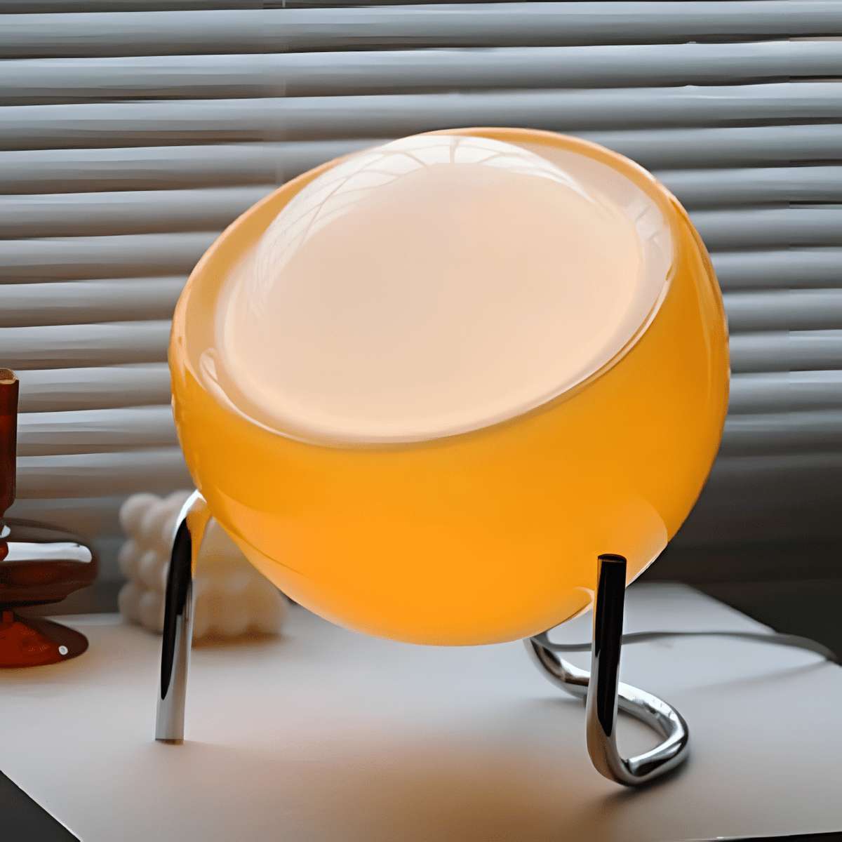 Bauhaus lampada da tavolo in vetro arancione e metallo cromato: primo piano del diffusore acceso sulla base cromata, davanti a una veneziana - Luximode