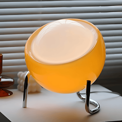 Bauhaus lampada da tavolo in vetro arancione e metallo cromato: primo piano del diffusore acceso sulla base cromata, davanti a una veneziana - Luximode