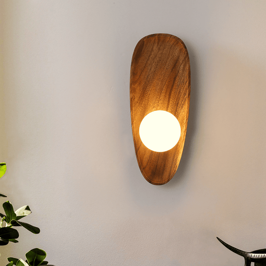 NaturalWooden applique da parete in legno con diffusore sferico bianco: applique accesa con luce calda su parete chiara accanto a una pianta - Luximode